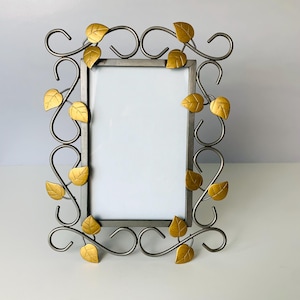 May include: A decorative metal picture frame with a rectangular opening. The frame is silver with gold-colored leaf accents. The leaves are evenly spaced around the frame, adding a natural touch to the design. The frame is designed to hold a photo or artwork.