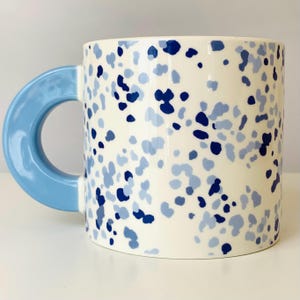 May include: A ceramic mug with a cream-colored base, adorned with blue and light blue speckled patterns. The mug features a large, rounded light blue handle. This coffee mug is a stylish addition to any kitchen.