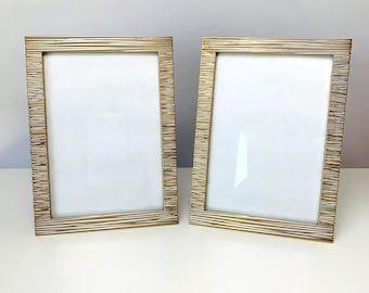 May include: Two gold-colored picture frames with a textured, horizontal line design. The frames have a rectangular shape with a white interior, suitable for displaying photos or artwork. The frames are displayed on a white surface.