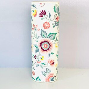 May include: A cylindrical vase with a white base featuring a repeating floral pattern in shades of pink, deep red, teal, and yellow. The design includes various stylized blossoms and leafy stems distributed across the entire exterior surface.