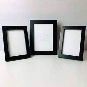 May include: Three black picture frames with white matting. The frames are rectangular with a simple, modern design. The center frame has the text "photo frame" and a decorative design. The frames are arranged on a white surface.