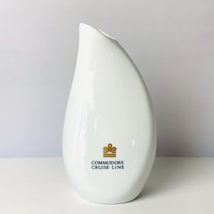 May include: White ceramic vase with a teardrop shape and a slanted opening. The vase features a gold logo that reads "COMMODORE CRUISE LINE". The vase has a glossy finish and is set against a white background.