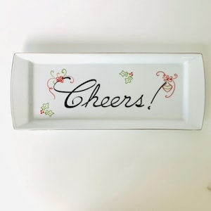 May include: White rectangular serving platter with a gold rim and the word "Cheers!" in black script. The platter is decorated with red and green holiday-themed accents, including holly and ornaments.