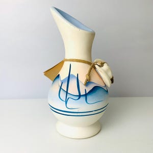 May include: A white ceramic pitcher with a curved spout and a round base. The pitcher is decorated with blue mountain designs and horizontal blue stripes. A small tag and a decorative hat are attached with twine.