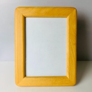 May include: A rectangular picture frame with rounded corners. The frame is made of light-colored wood with a warm, yellow hue. The frame surrounds a white interior, ready for a photograph or artwork. The frame is designed to stand on a flat surface.