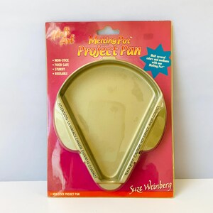 May include: A gold-colored non-stick Melting Pot Project Pan on a red and pink background. The package includes text that says "Non-Stick", "Food Safe", "Sturdy", and "Reusable". The pan is designed to melt several colors and mediums.