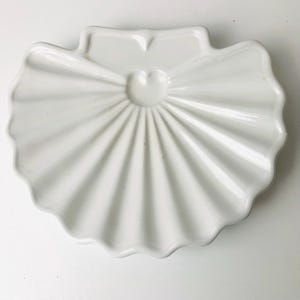 May include: A white ceramic seashell-shaped dish with a scalloped edge. The dish has a central, raised circular design and radiating lines, resembling a seashell. The dish is a decorative item.