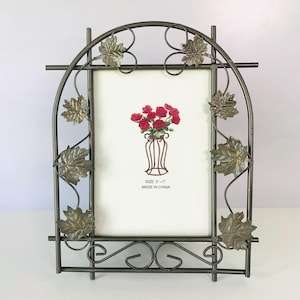 May include: A dark gray metal picture frame with an arched top and decorative metal maple leaves attached to the sides. The frame holds a white insert featuring a red rose bouquet illustration, the text SIZE: 5" x 7", and MADE IN CHINA.