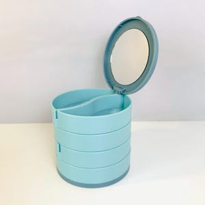 May include: A light blue, cylindrical, multi-tiered storage organizer with four rotating compartments. The top lid features an integrated circular mirror on the underside, and the interior is divided into two sections for organizing small items.