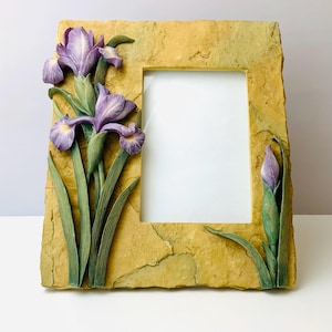 May include: A decorative photo frame with a rectangular opening. The frame is a textured, tan color with a sculpted design of purple irises and green leaves. The frame is made of a stone-like material.