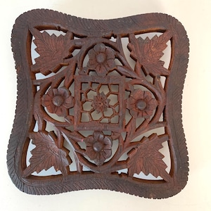 May include: A square, dark brown wooden trivet with intricate floral and leaf carvings. The trivet features a central square with a geometric design, surrounded by flowers and leaves. The edges have a decorative, textured border.