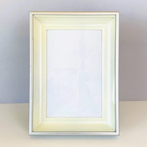 May include: A rectangular picture frame featuring a metallic silver outer border and a wide, pale yellow inner beveled frame. The center contains a blank white rectangular insert, and the entire structure is oriented vertically for display.