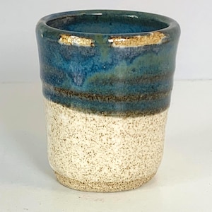May include: Handmade ceramic tumbler with a speckled off-white base, a dark brown band, and a gradient of blue and teal at the top. The rim has a gold-colored accent. The cup is cylindrical and has a rustic, artisanal appearance.