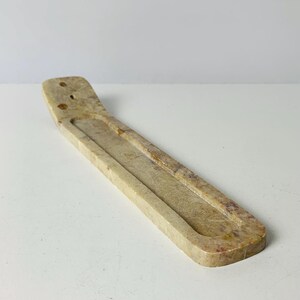 May include: A beige incense holder carved from stone. The rectangular holder has a long, shallow groove for incense sticks and two small holes at one end. The stone has natural variations in color, with hints of brown and tan.