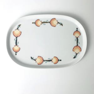 May include: White oval serving platter with a seashell design. The platter features a border of peach-colored seashells and teal-colored vines. The seashells are arranged in a repeating pattern around the edge of the platter.