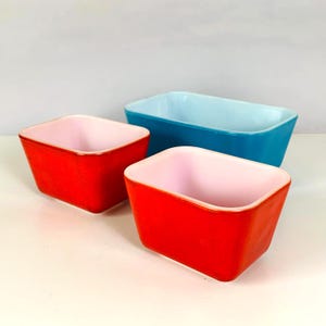 May include: A set of three rectangular baking dishes with rounded corners. Two dishes feature a vibrant red exterior with a pale pink interior, while the third, larger dish has a bright turquoise exterior with a light blue interior finish.