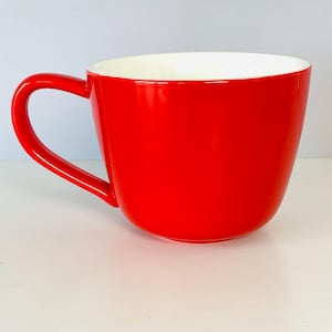 May include: A bright red ceramic mug with a smooth, glossy finish and a white interior. The mug features a wide, rounded body and a curved handle on the left side, providing a simple and functional design for holding hot or cold beverages.