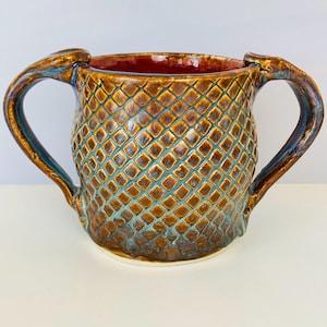 May include: A handcrafted ceramic mug with two handles. The mug features a textured, diamond-patterned exterior in shades of brown and teal. The interior is a deep red color, and the handles have a curved design. This unique mug is perfect for coffee or tea.