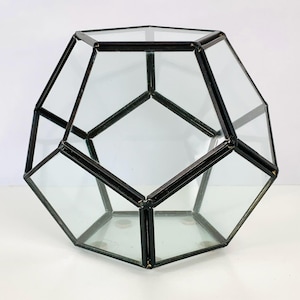 May include: A geometric terrarium featuring a dodecahedron shape constructed from clear glass panels held together by a dark black metal frame. The structure has multiple pentagonal faces and is designed as a hollow, multi-faceted container.