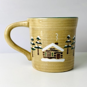 May include: A tan ceramic mug with a curved handle. The mug features a winter scene with a cabin, snow, and green trees. The rim of the mug is green.