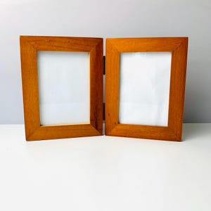 May include: A hinged, double-frame picture frame made of stained wood. The frames are rectangular with a light brown finish and a white interior. The two frames are connected by a hinge, allowing them to stand upright or fold flat.