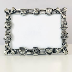 May include: A rectangular silver-toned picture frame featuring a raised relief design of various teacups and saucers along the border. The four corners are decorated with sculpted bows, and the frame has a horizontal ribbed texture throughout.