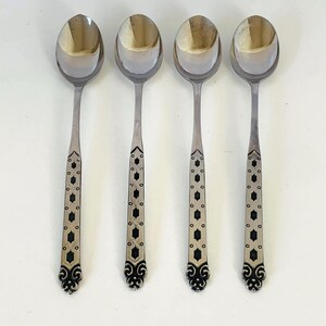 May include: Four silver-toned metal spoons arranged in a vertical row. Each spoon features a polished oval bowl and a long, slender handle decorated with a repeating black geometric pattern of diamonds and dots, ending in a decorative scroll motif.