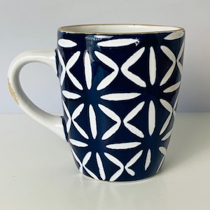 May include: A ceramic mug with a navy blue background and a white geometric pattern. The mug has a white handle and a gold rim. The handle has a distressed look. The mug is designed for hot beverages.
