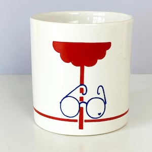 May include: A white ceramic mug featuring a graphic design of a red scalloped shape atop a vertical red line, intersected by a horizontal red line. Centered over the red lines is a blue line drawing of round eyeglasses with a small heart detail.