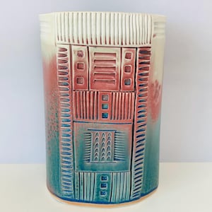 May include: A cylindrical ceramic vase featuring a gradient of teal and reddish-pink glaze. The surface is decorated with carved geometric patterns, including vertical lines, squares, and rectangular blocks, creating a textured relief design.