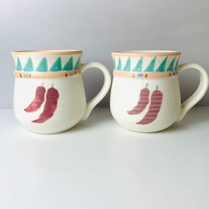 May include: Two off-white ceramic mugs with a rounded shape and a handle. Each mug features a design of two red chili peppers and a decorative border with teal triangles, peach, and blue accents. The mugs are suitable for hot or cold beverages.