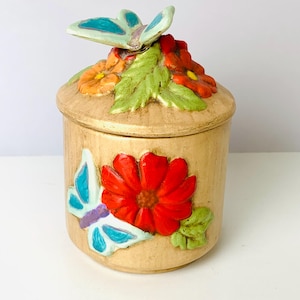 May include: A tan ceramic jar with a lid, decorated with a red flower and a blue butterfly. The lid features a butterfly and flowers. The jar is cylindrical and has a textured surface.