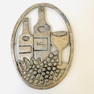 May include: Oval-shaped, silver-colored metal trivet featuring a wine theme. The design includes two wine bottles, a wine glass, and a cluster of grapes. The word "Vine" is written on one of the bottles.