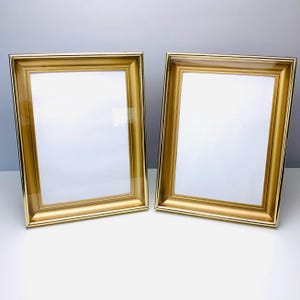 May include: Two gold-colored picture frames with a rectangular shape. The frames have a glossy finish and a simple, elegant design. The frames are empty, with a white background visible through the glass.