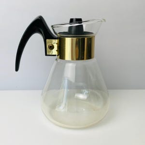May include: A clear glass carafe with a black handle and a gold-colored band around the middle. The carafe has a spout for pouring and a black lid. The design is vintage and elegant, suitable for serving beverages.
