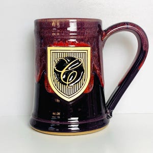 May include: A handcrafted ceramic mug with a burgundy glaze and a large handle. The mug features a crest design with a gold border and a black interior with a stylized letter "C". The mug is suitable for hot or cold beverages.
