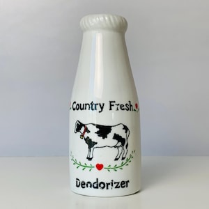 May include: White ceramic milk bottle-shaped deodorizer with a black and white cow illustration. The bottle has the words "Country Fresh" and "Dendorizer" printed on it, with a red heart and green leaves.