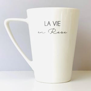 May include: A white ceramic mug with a curved handle. The mug features the phrase "LA VIE en Rose" in black script. The mug has a smooth, glossy finish and a simple, elegant design, suitable for coffee or tea.