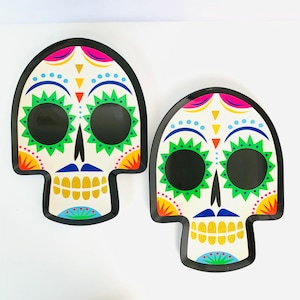 May include: Two skull-shaped serving trays with a white base and black borders. Each tray features a colorful graphic design with large black eye sockets, green starburst patterns, yellow teeth, and accents in blue, pink, orange, and yellow.