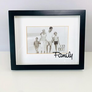 May include: Black square picture frame with a white mat and a family photo. The photo shows a family of four on a beach. The word "Family" is written in cursive on the frame.