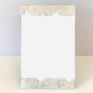 May include: A rectangular vertical picture frame featuring a plain white center panel. The top and bottom edges are adorned with a raised, metallic silver-toned border displaying an intricate, embossed scrolling vine and leaf pattern.