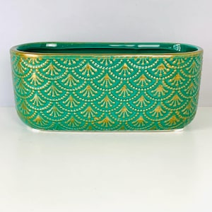 May include: An oval-shaped ceramic planter featuring a vibrant teal green base color with a repeating embossed fan-like pattern highlighted in metallic gold. The rim of the planter is also finished with a thin, consistent metallic gold trim.