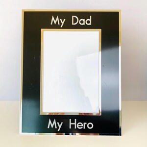 May include: A rectangular black picture frame with a gold border and the words "My Dad" and "My Hero" in white lettering. The frame has a white interior and is designed to hold a photograph.