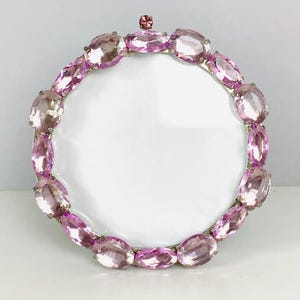 May include: A round silver picture frame with a border of pink oval-shaped gemstones. A smaller pink gemstone sits at the top center. The frame has a white interior.