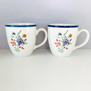 May include: Two white ceramic mugs with a blue rim and a floral design. The floral design features red, blue, and yellow flowers with green leaves. The mugs have a curved handle.