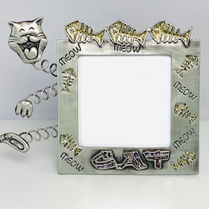 May include: A silver-colored cat-themed picture frame with the word "CAT" in colorful letters at the bottom. The frame has a cat head and tail design, with "meow" written around the frame and fish bone accents.