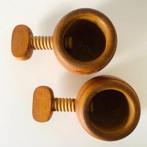 May include: Two round, light brown wooden nutcrackers with a screw-style mechanism. Each tool features a cylindrical body with a hollow center and a T-shaped handle attached to a threaded screw that adjusts the internal space for cracking nuts.