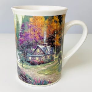 May include: White ceramic mug featuring a scenic landscape with a stone cottage, surrounded by colorful autumn trees. The mug has a curved handle and a wide rim. The artwork depicts a path leading to the cottage, with a stream and flowers.
