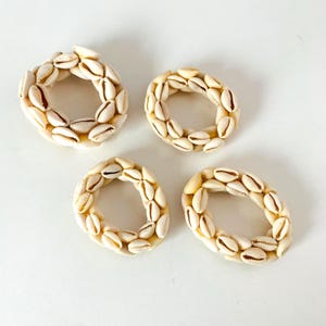 May include: Four decorative napkin rings. Each ring is made of small, cream-colored seashells arranged in a circular pattern. The shells have a natural, organic look, with a mix of cream and tan hues. The rings are set against a white background.