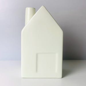 May include: A cream-colored, house-shaped ceramic vase with a chimney and a rectangular window detail. The vase is a minimalist design, perfect for home decor. The house is approximately 8 inches tall.
